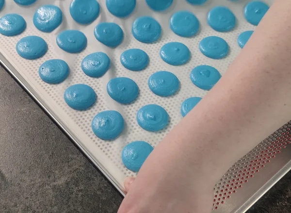 Handen houden een bakplaat met versgebakken blauwe macaronschelpen vast