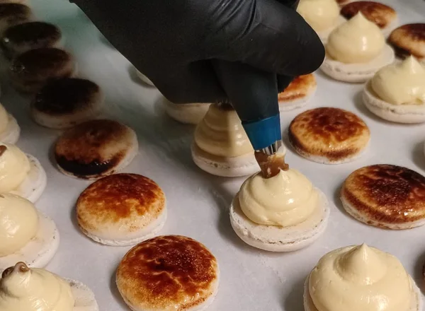 Een gehandschoende hand spuit de vulling op macarons in het atelier